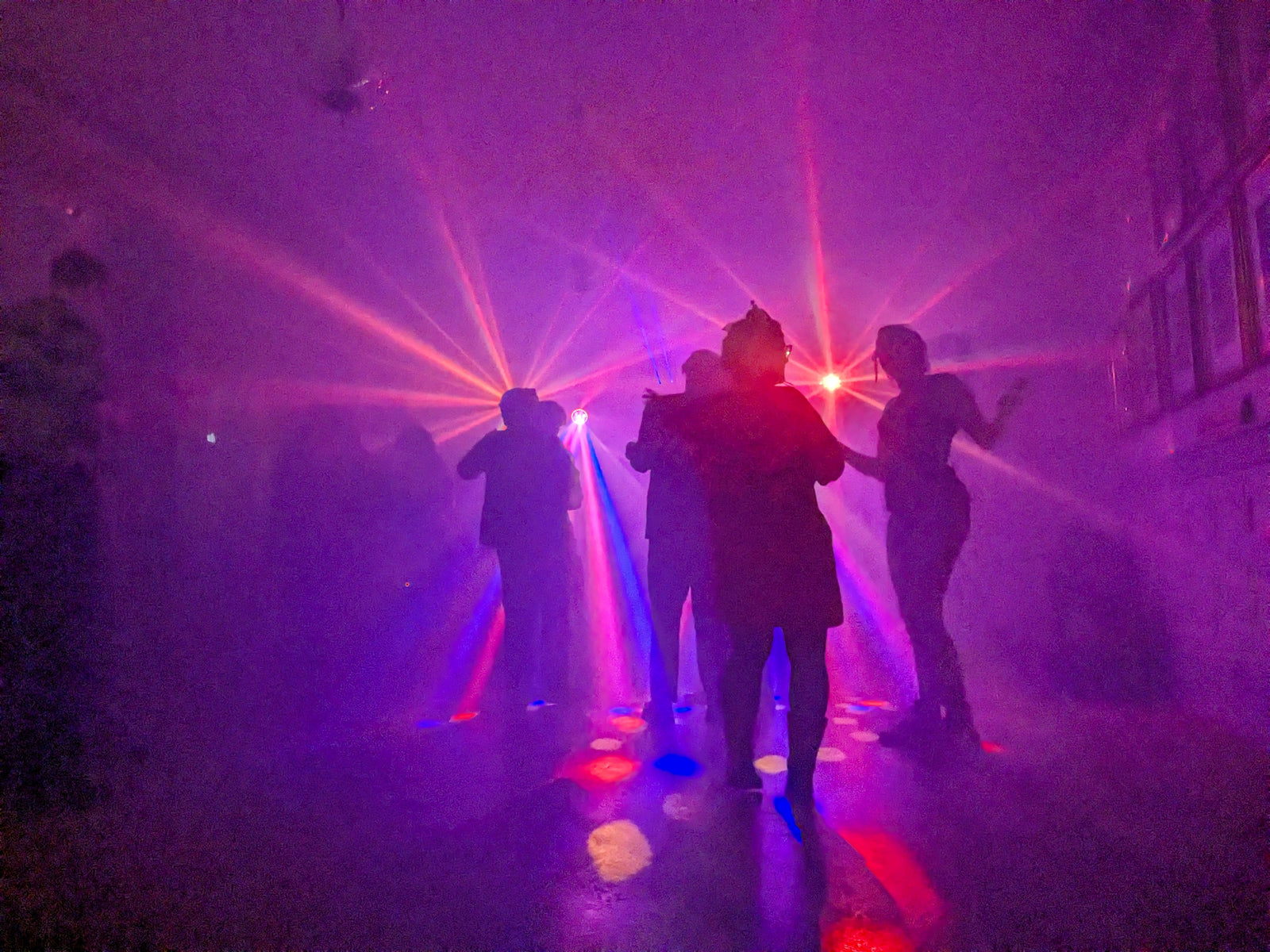 Personer dansar i färgglada ljusstrålar från laser- och discoljus i ett rum fyllt med rökeffekt, vilket skapar en livlig feststämning med lila och röda toner.