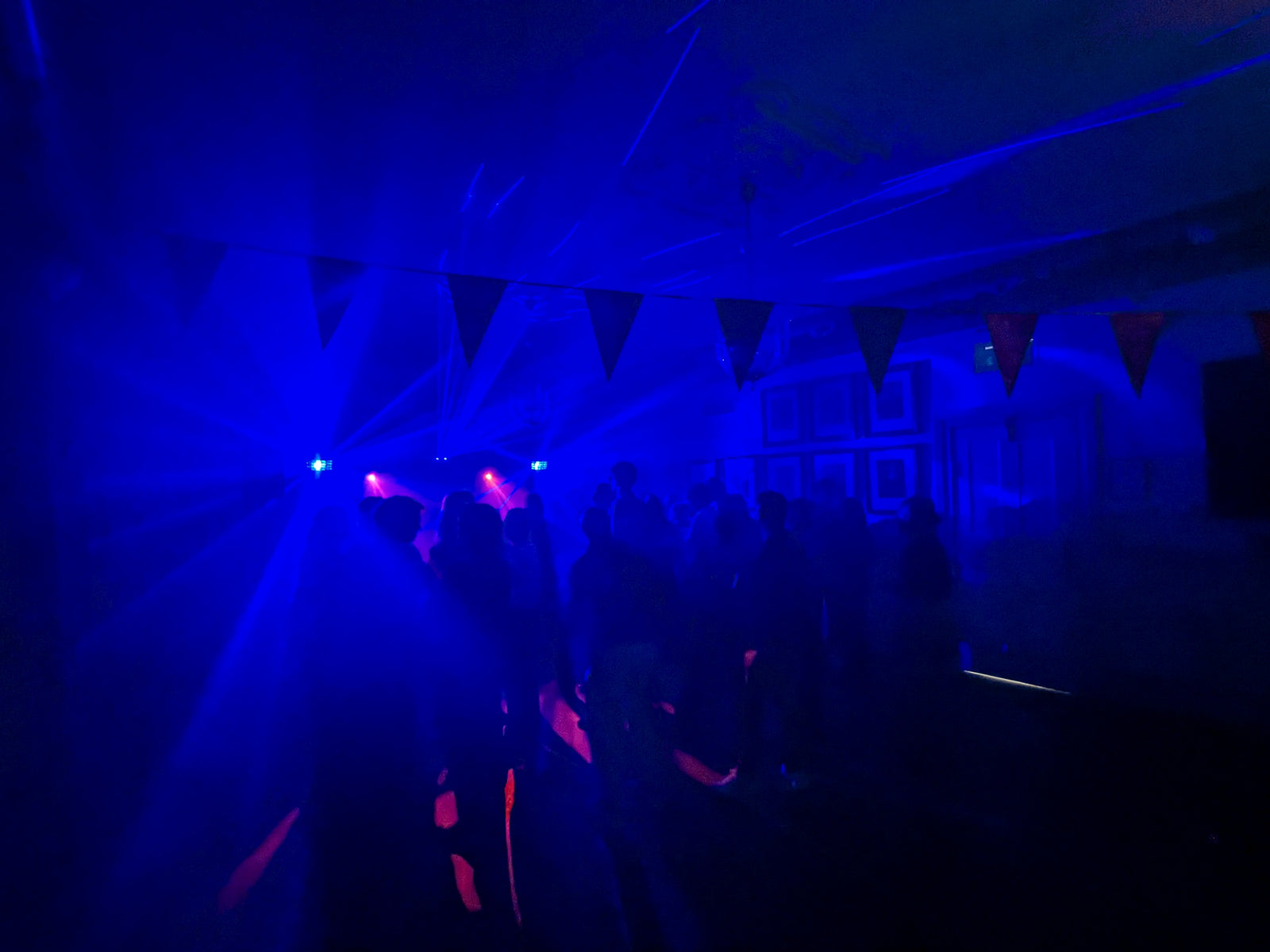 Dansgolv fyllt med människor under blå och lila ljuseffekter, med rökeffekt och laserstrålar som sprids i rummet dekorerat med vimplar i taket.