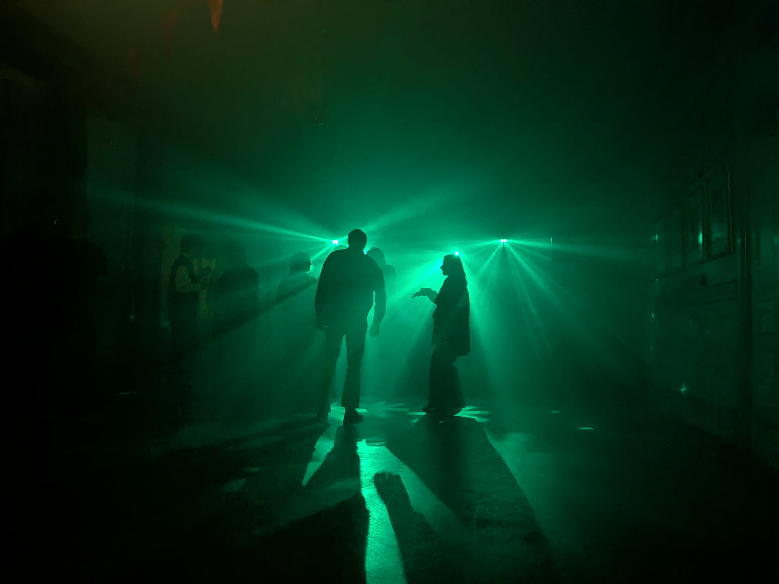 Silhuetter av personer som dansar i grönt laserljus och rökeffekt, där ljusstrålarna sprider sig dramatiskt över dansgolvet i ett mörkt rum.