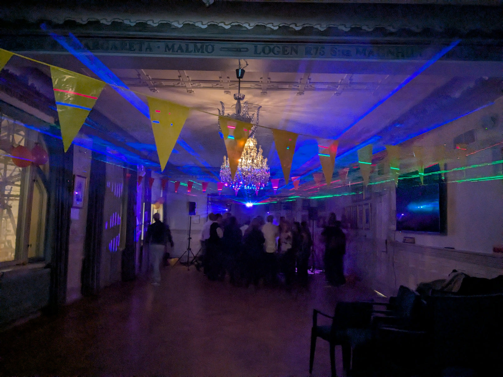 Festlokal dekorerad med färgglada vimplar och kristallkrona, där laser- och discoljus i grönt och lila lyser upp rummet medan människor dansar längst bak.