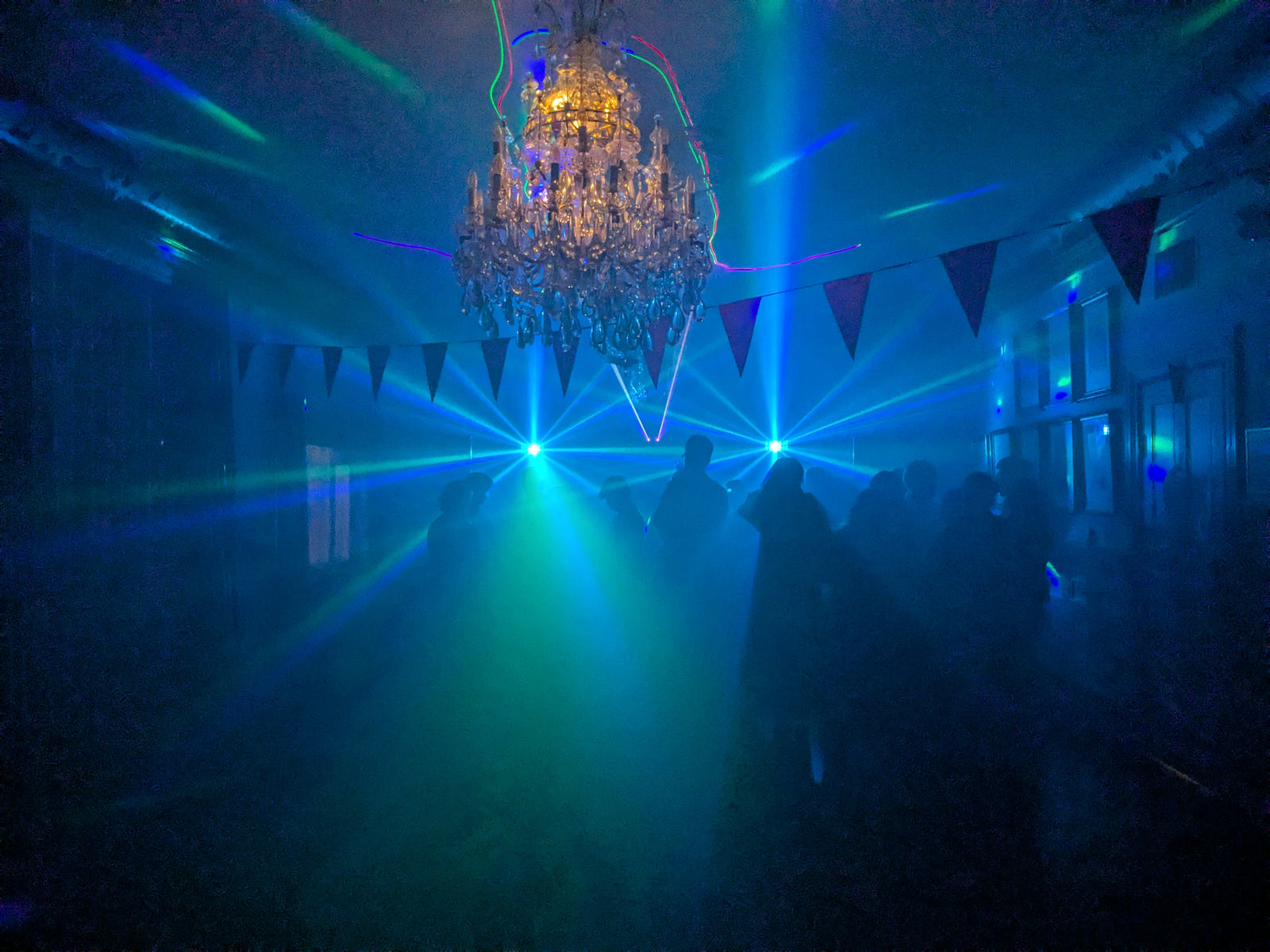 Dansgolv upplyst av blå laserstrålar under en kristallkrona, med människor som dansar i rummet dekorerat med vimplar och fyllt med rökeffekt.
