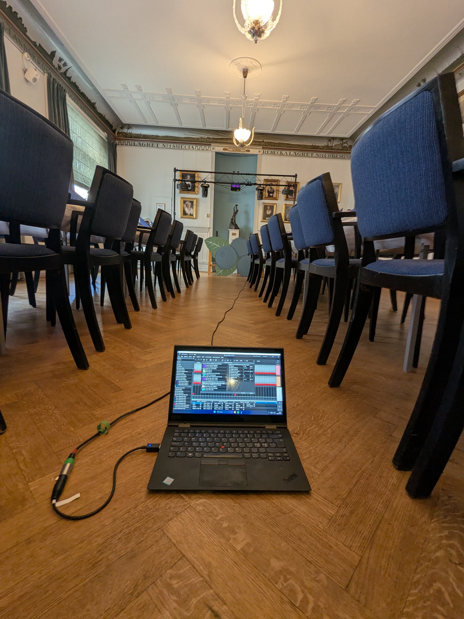 Laptop placerad på ett trägolv mellan två rader stolar i en festlokal, ansluten med kablar och visande ljusprogramvara för scenbelysning uppsatt längst fram i rummet.