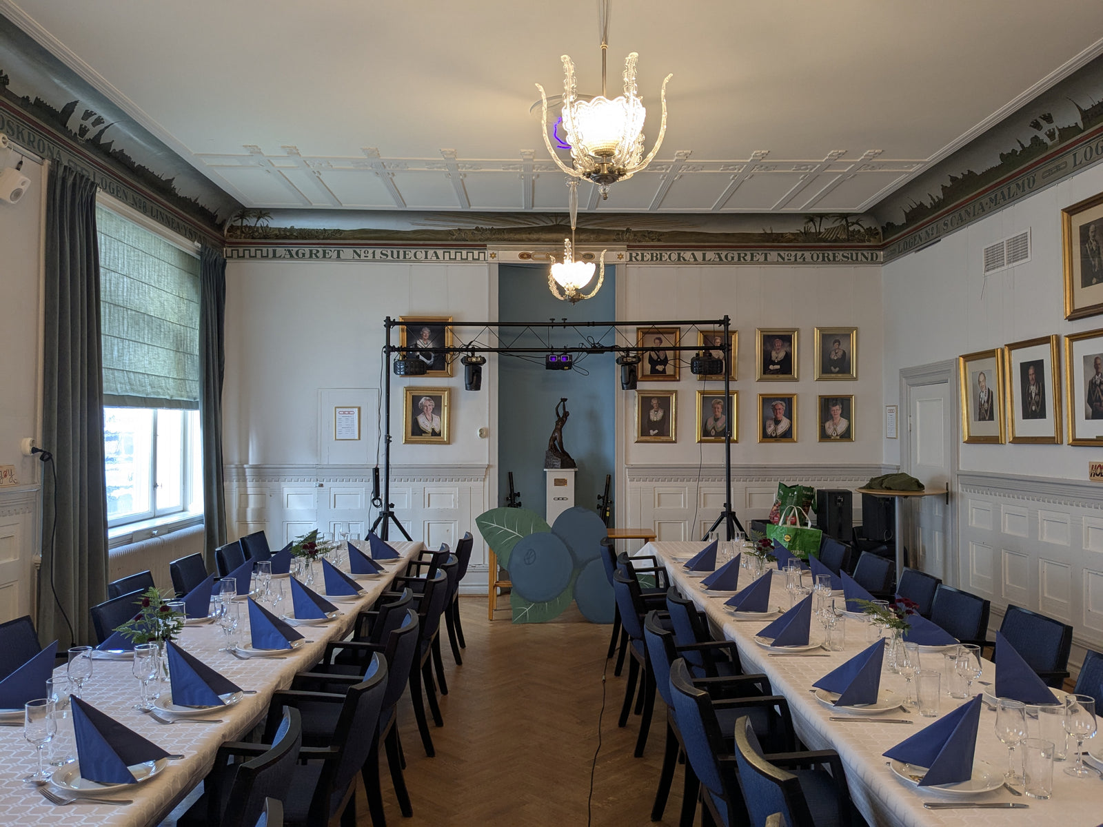 "Ljusstativ med upphängda scenlampor och ljuseffekter uppställt längst fram i en dukad festlokal med långbord, blå servetter och porträtttavlor på väggarna.